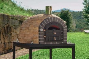 Un four à bois extérieur traditionnel en briques de terre cuite avec une porte en fer forgé, posé sur un support métallique noir devant un mur de pierre et une colline verdoyante