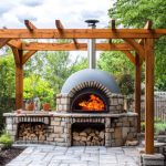 Un four à bois extérieur en pierre et brique, allumé avec de grandes flammes, installé sous une pergola en bois massif sur une terrasse pavée dans un jardin paysager.
