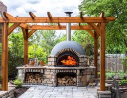 Un four à bois extérieur en pierre et brique, allumé avec de grandes flammes, installé sous une pergola en bois massif sur une terrasse pavée dans un jardin paysager.