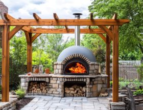 Un four à bois extérieur en pierre et brique, allumé avec de grandes flammes, installé sous une pergola en bois massif sur une terrasse pavée dans un jardin paysager.