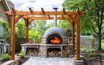Un four à bois extérieur en pierre et brique, allumé avec de grandes flammes, installé sous une pergola en bois massif sur une terrasse pavée dans un jardin paysager.