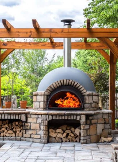 Un four à bois extérieur en pierre et brique, allumé avec de grandes flammes, installé sous une pergola en bois massif sur une terrasse pavée dans un jardin paysager.