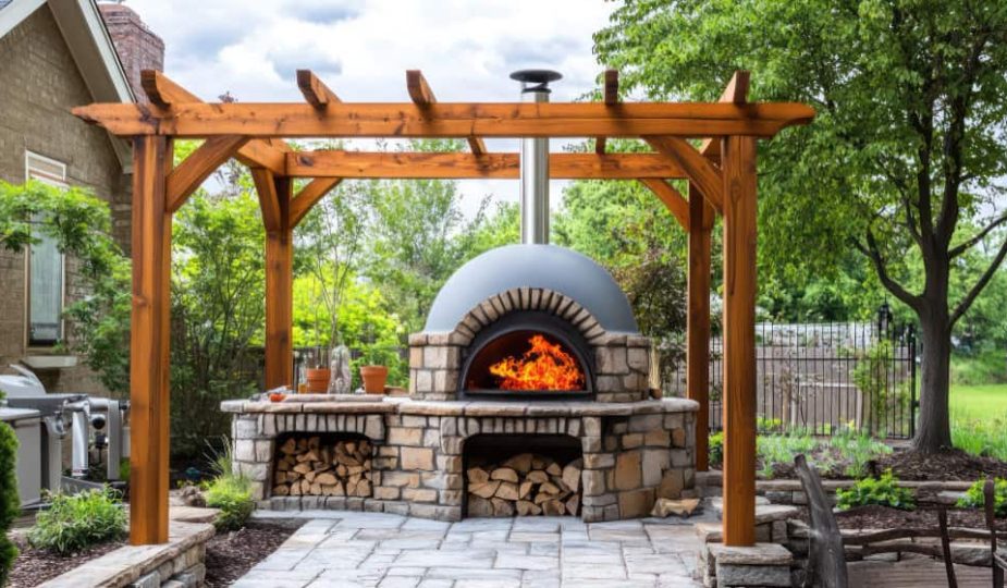 Un four à bois extérieur en pierre et brique, allumé avec de grandes flammes, installé sous une pergola en bois massif sur une terrasse pavée dans un jardin paysager.